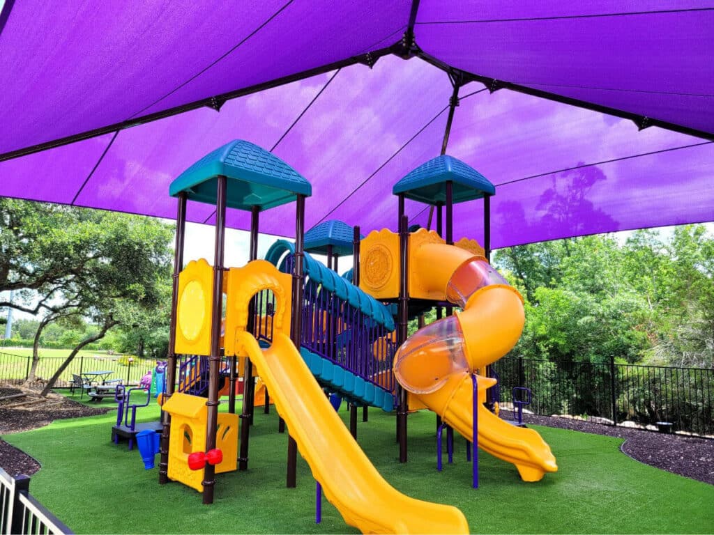 Commercial playground installed on turf under a hip shade structure in Austin, Texas, designed for safety, comfort, and year-round outdoor play.
